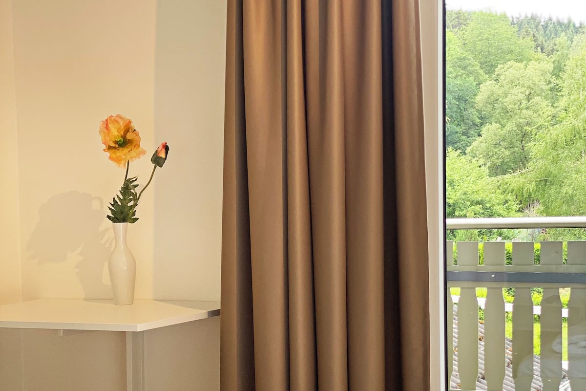 A bedroom with a white bed, a white nightstand, a vase with a single flower, and a view of a balcony with a white fence and greenery outside.