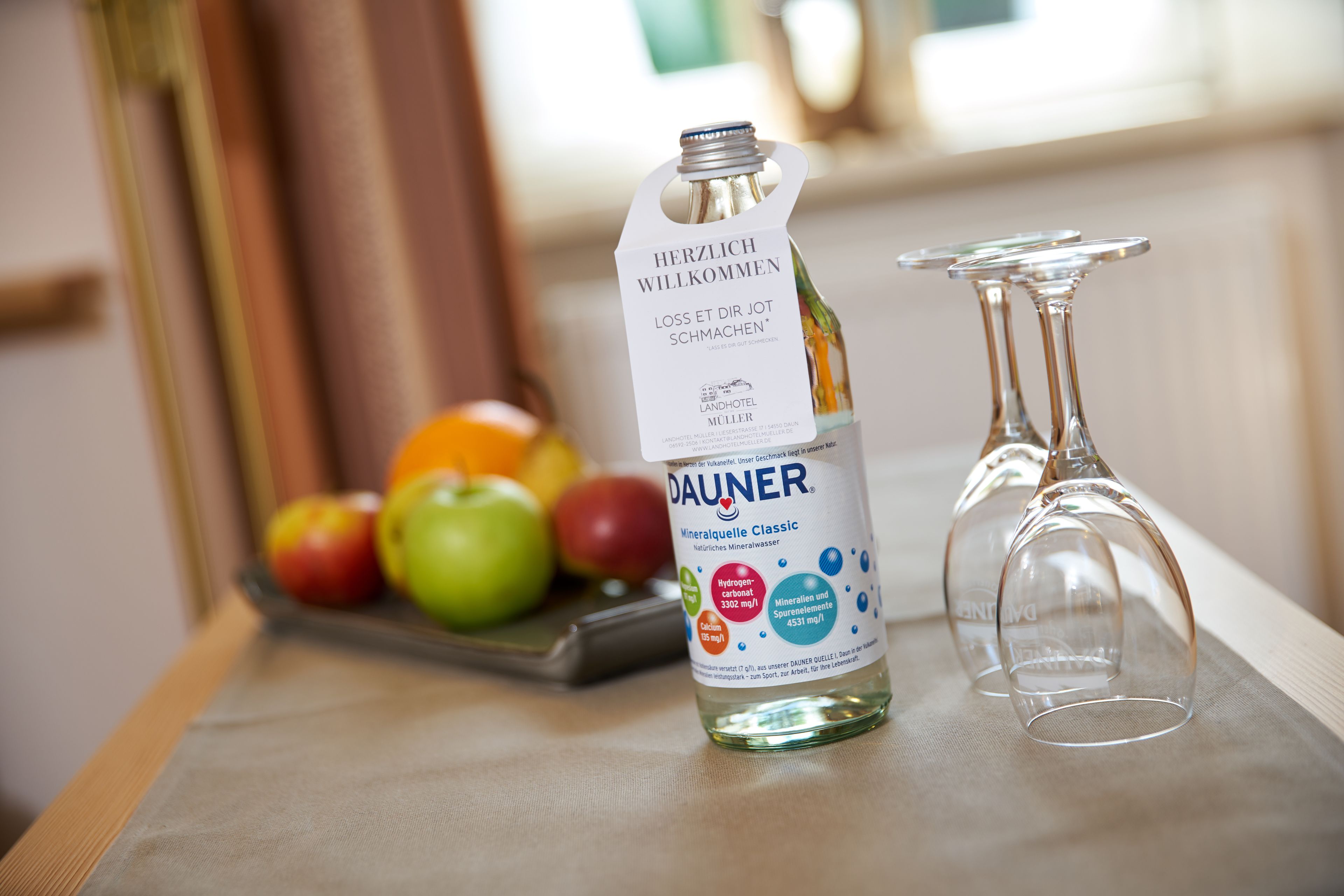 Une bouteille d’eau minérale DAUNER avec une étiquette, une assiette de fruits et deux verres à vin vides sur une table.