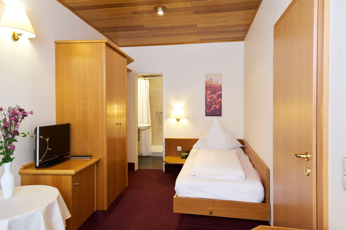 A hotel room with a wooden bed, a TV on a wooden stand, a bedside table with a lamp, and a bathroom visible through an open door.