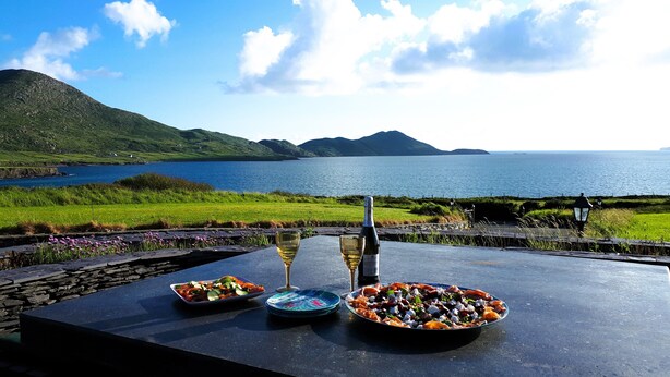 Ein Tisch mit Speisen und einer Flasche, vor einer malerischen Kulisse aus See und Bergen.