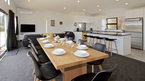 Una cocina moderna con una isla grande, un comedor con una mesa larga dispuesta para seis personas, y una sala de estar con sofá y televisor.