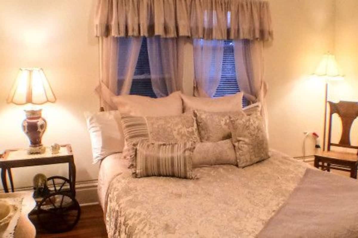 Un dormitorio con cama, mesitas de noche, un espejo, una ventana con cortinas, un aparador y una silla.