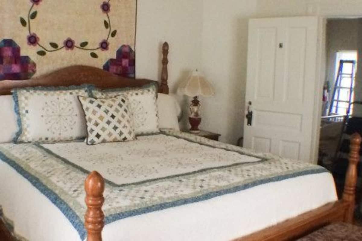 Una cama bien tendida con un cabecero de madera, dos mesitas de noche con lámparas y un tapiz floral en la pared.