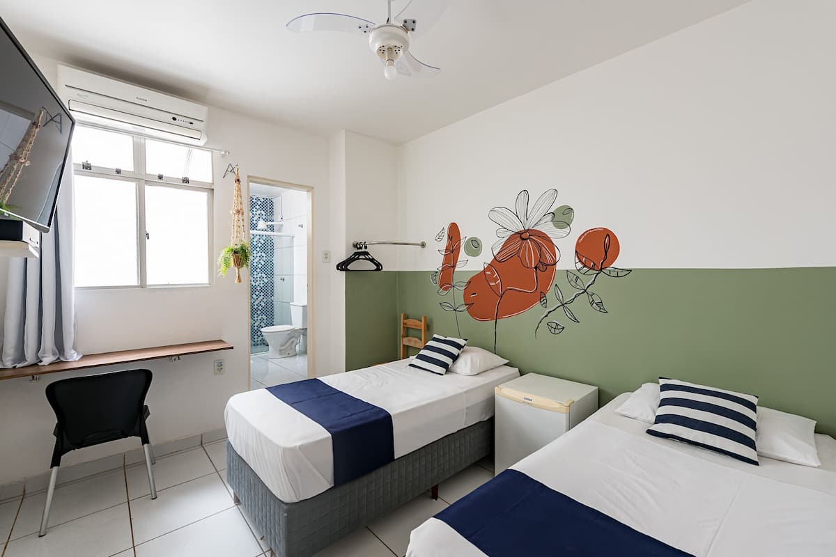 A hotel room with two beds, a desk, a chair, a ceiling fan, and a window.