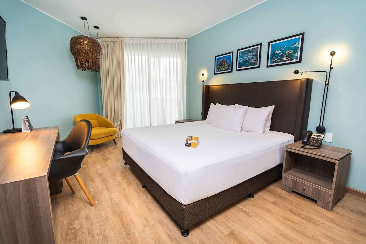 A hotel room with a bed, a desk, a chair, a nightstand, and a lamp.