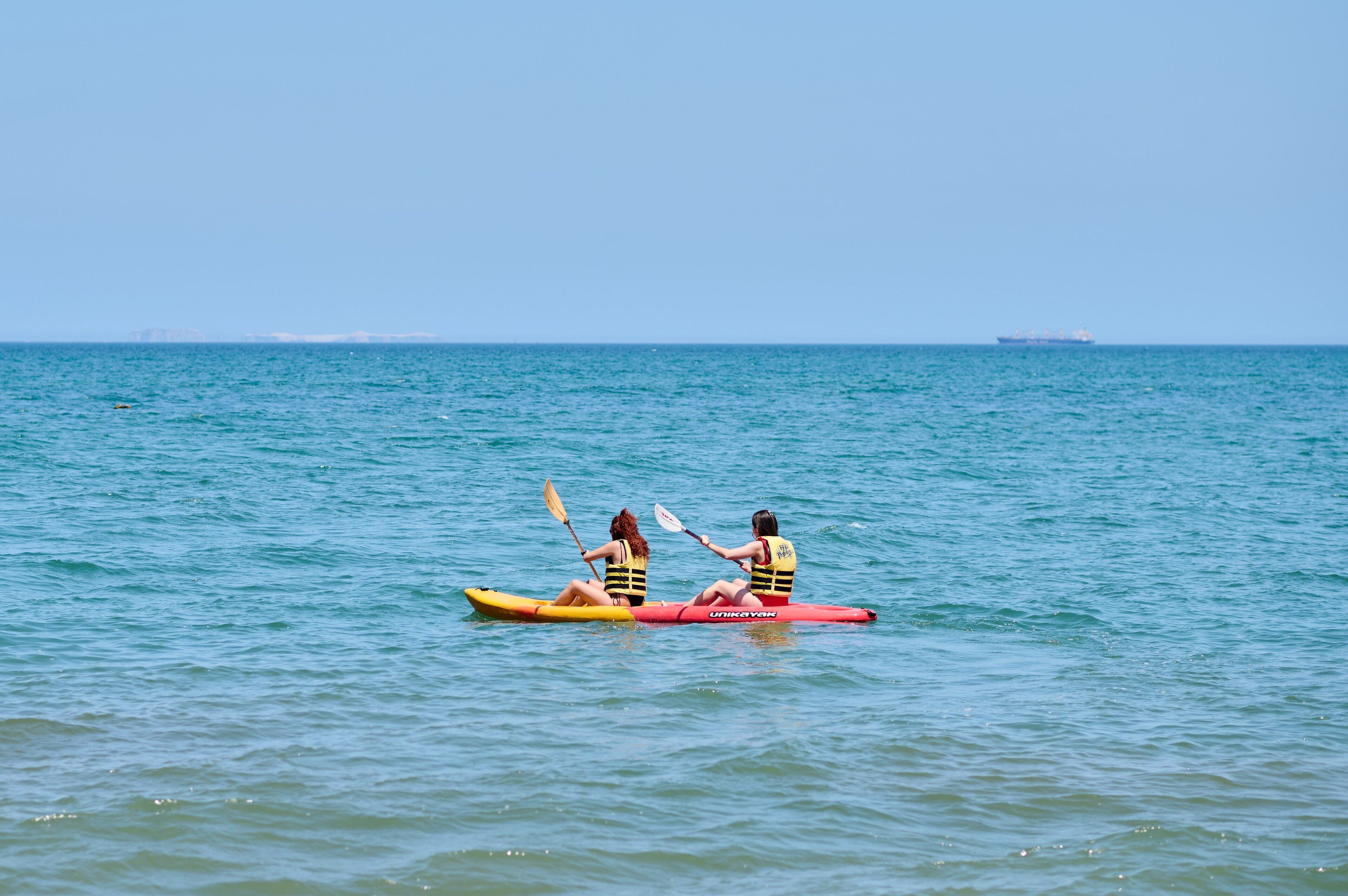 Dos personas kayakando en un mar tranquilo, con un barco distante a la vista.