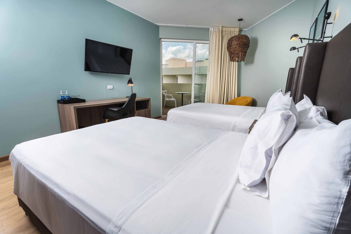 A hotel room with two beds, a desk, a chair, a TV, and a balcony with a view of the sky.