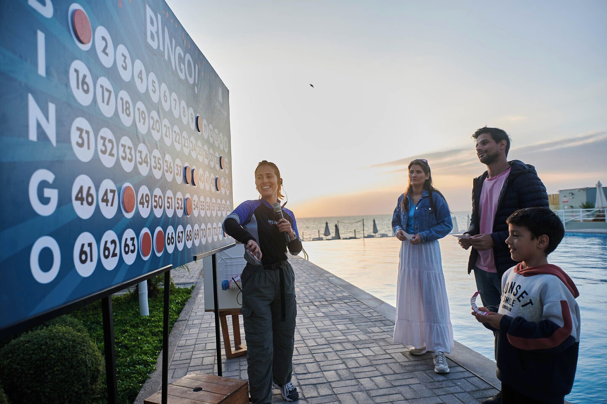 Un grupo de personas de pie cerca de un gran cartel con números y letras, posiblemente para un juego o evento, junto a la orilla del agua.