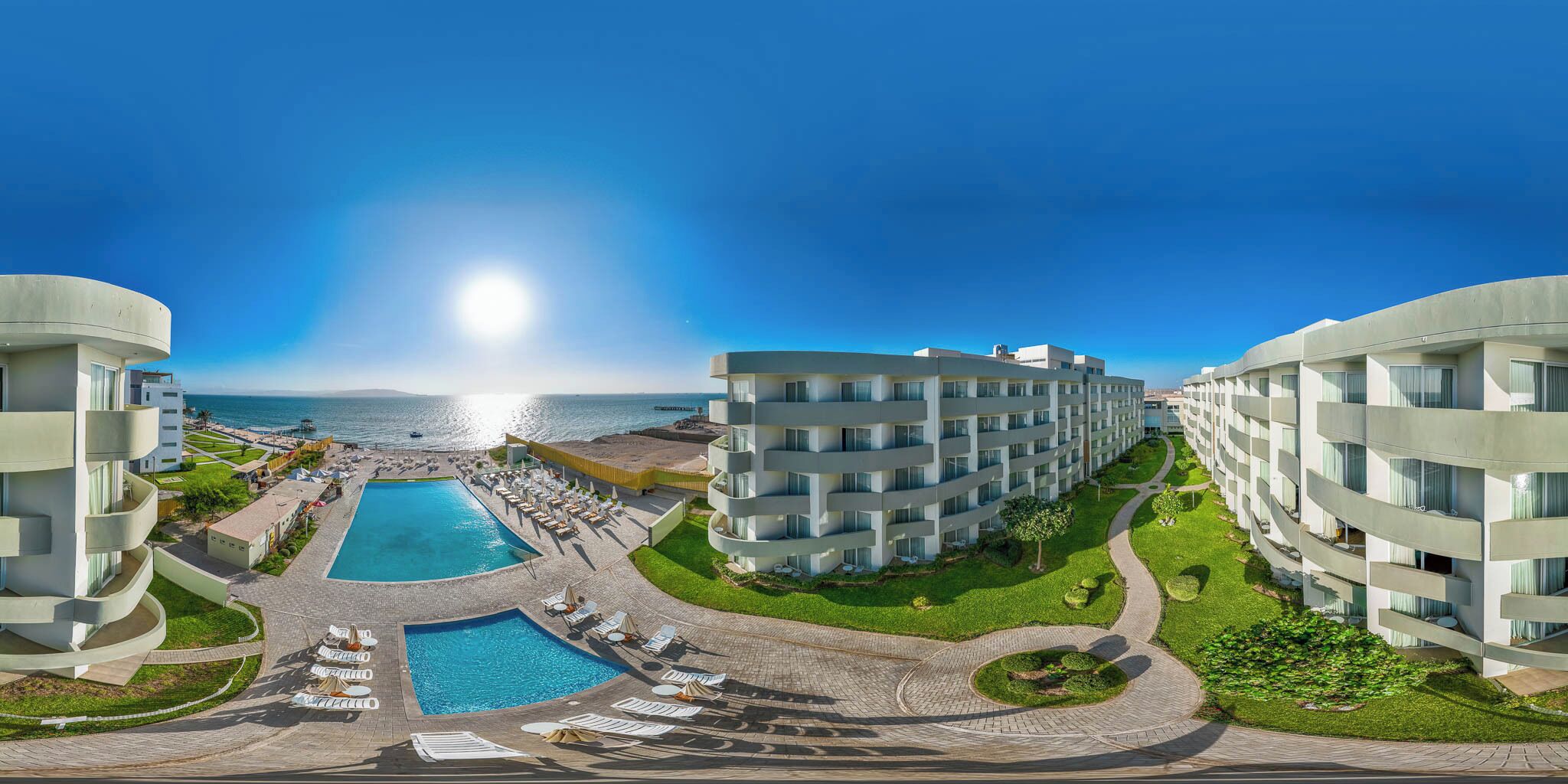 A resort with a central pool, multiple buildings, and a view of the sea.