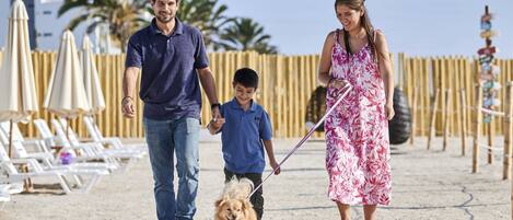 A family walking a dog on a sandy path with beach umbrellas and lounge chairs in the background.