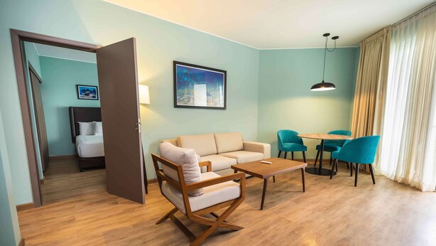 Une chambre d’hôtel moderne avec un plancher en bois, un canapé beige, une table basse en bois et des chaises turquoise disposées autour d’une petite table. On aperçoit un lit avec une literie blanche à l’arrière-plan.