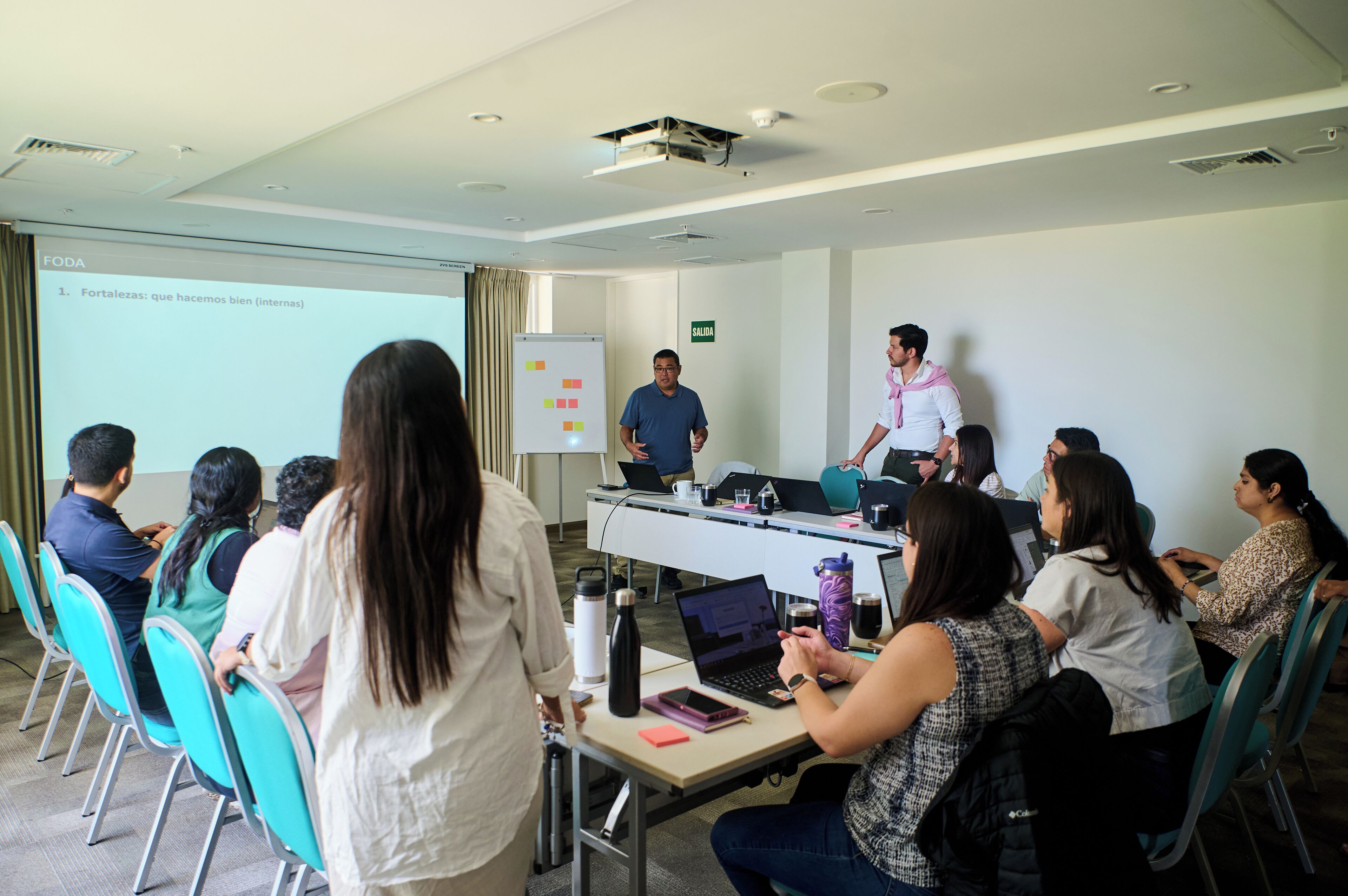 A conference room with a presentation screen, a whiteboard, and attendees seated at tables with laptops and notebooks.