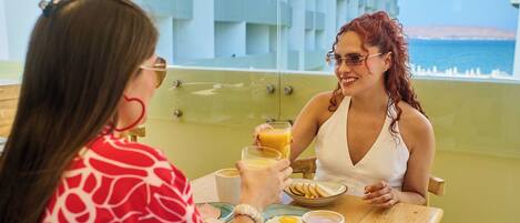 Dos personas sentadas a una mesa con alimentos para el desayuno, como huevos y jugo de naranja, en una habitación con vista al mar.