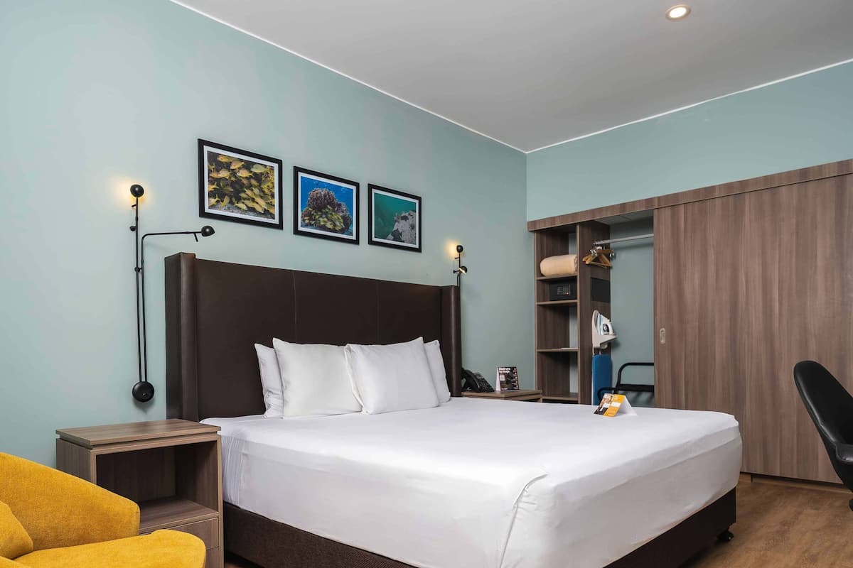 A modern hotel room with a large bed, a wooden floor, a yellow armchair, a wardrobe, and a nightstand.