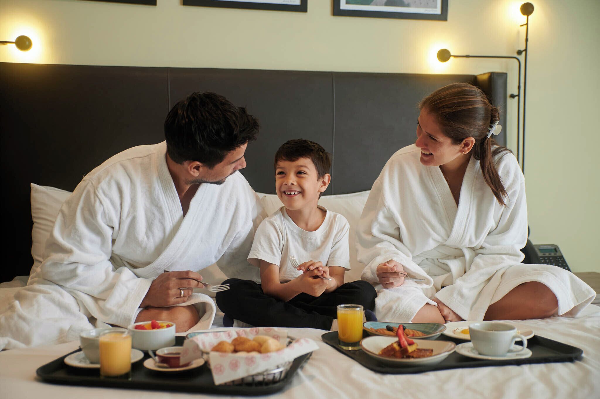 A family of four in a hotel room with a bed, breakfast tray, and a bedside table.