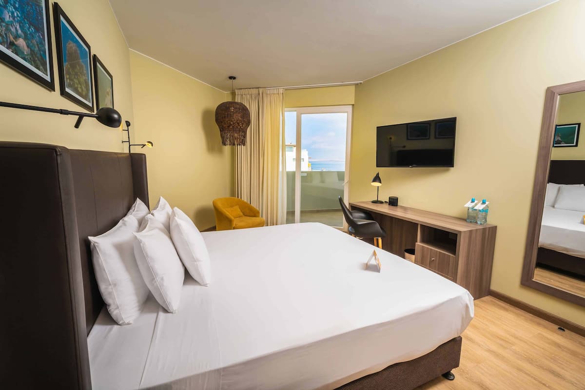 A hotel room with a large bed, a desk, a chair, a TV, and a balcony with a view.