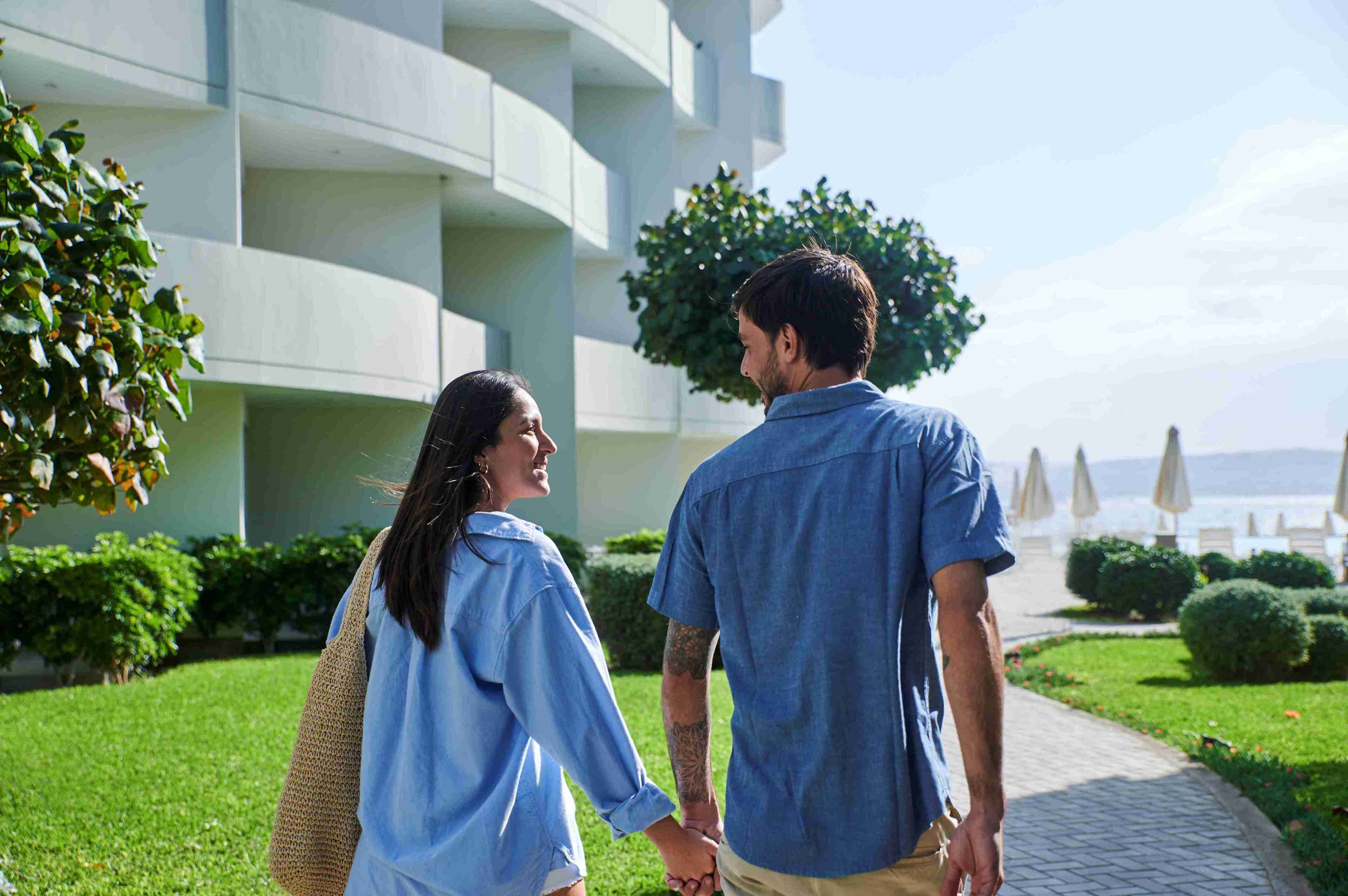 Um casal caminhando por um caminho, com um edifício moderno e vista para o mar ao fundo.
