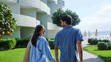 Um casal caminhando por um caminho, com um edifício moderno e vista para o mar ao fundo.