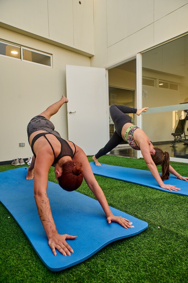 Dos personas practicando yoga sobre esterillas azules en una habitación con una puerta blanca y un muro de cristal.