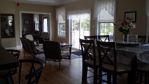 A dining area with a table set for four, surrounded by chairs. There is a window with white curtains, a door leading to another room, and a small table with a vase of flowers.