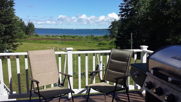A deck with two chairs and a grill, overlooking a grassy area and a body of water.
