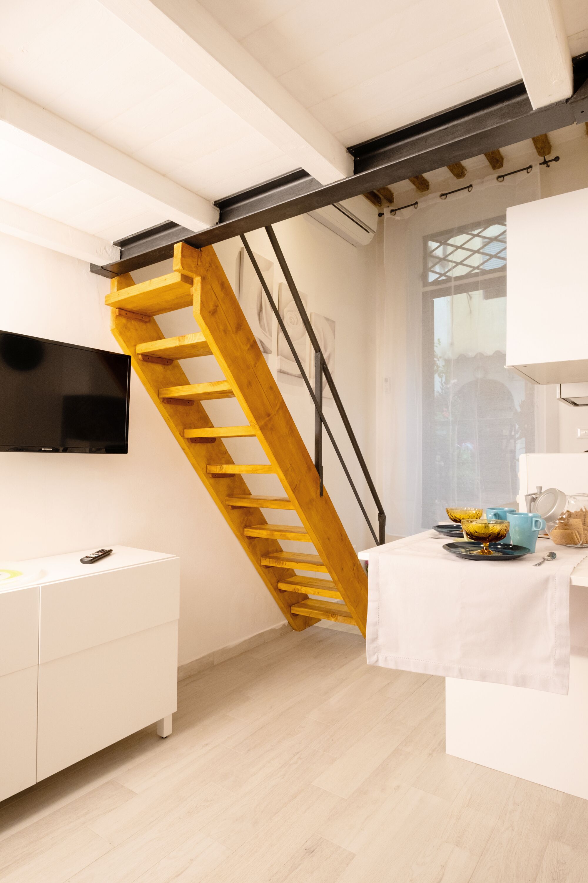Un interior modern amb una escala de fusta, un televisor de pantalla plana i una zona de menjador amb una taula preparada per a dues persones.