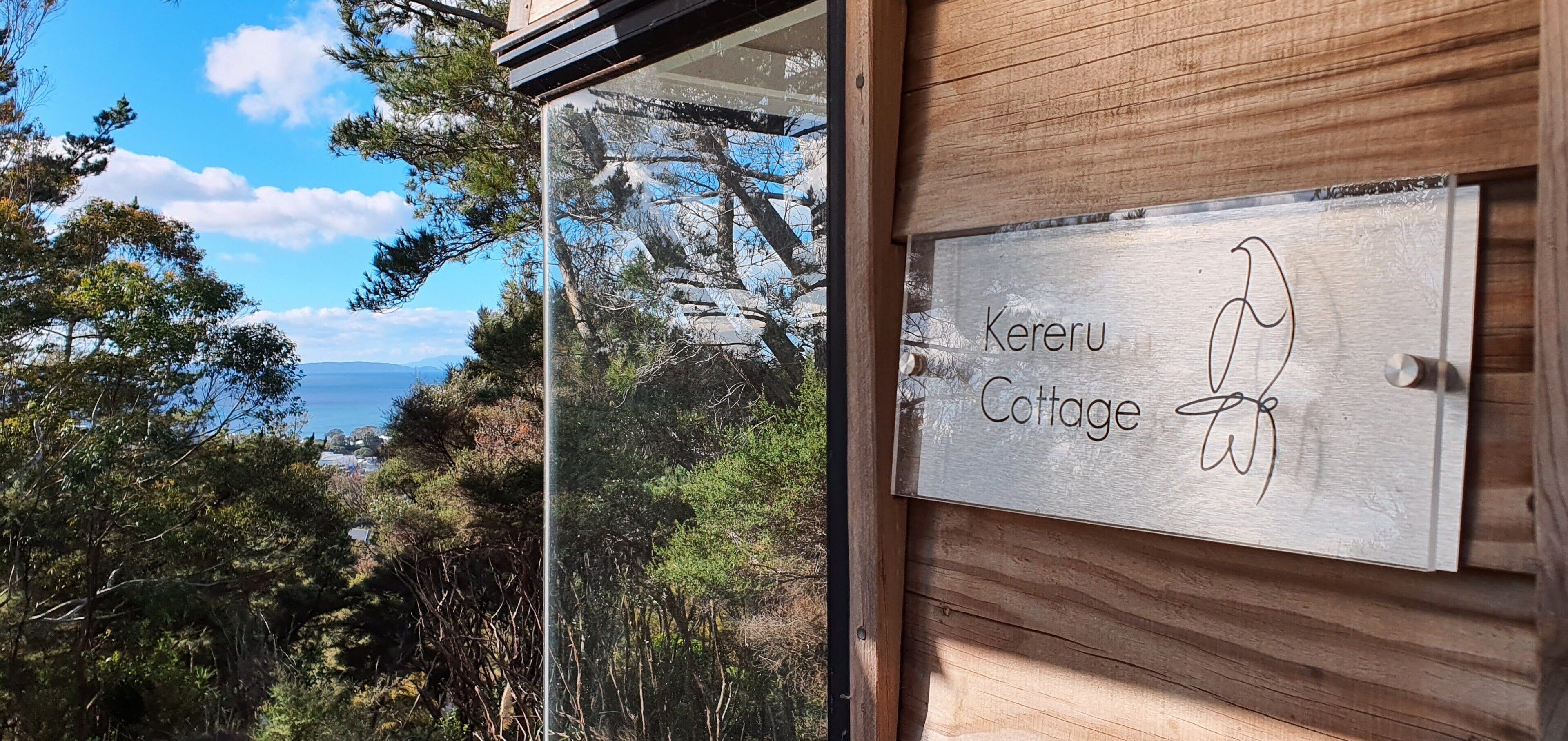A wooden sign reading "Kereru Cottage" with a view of trees and mountains in the background.