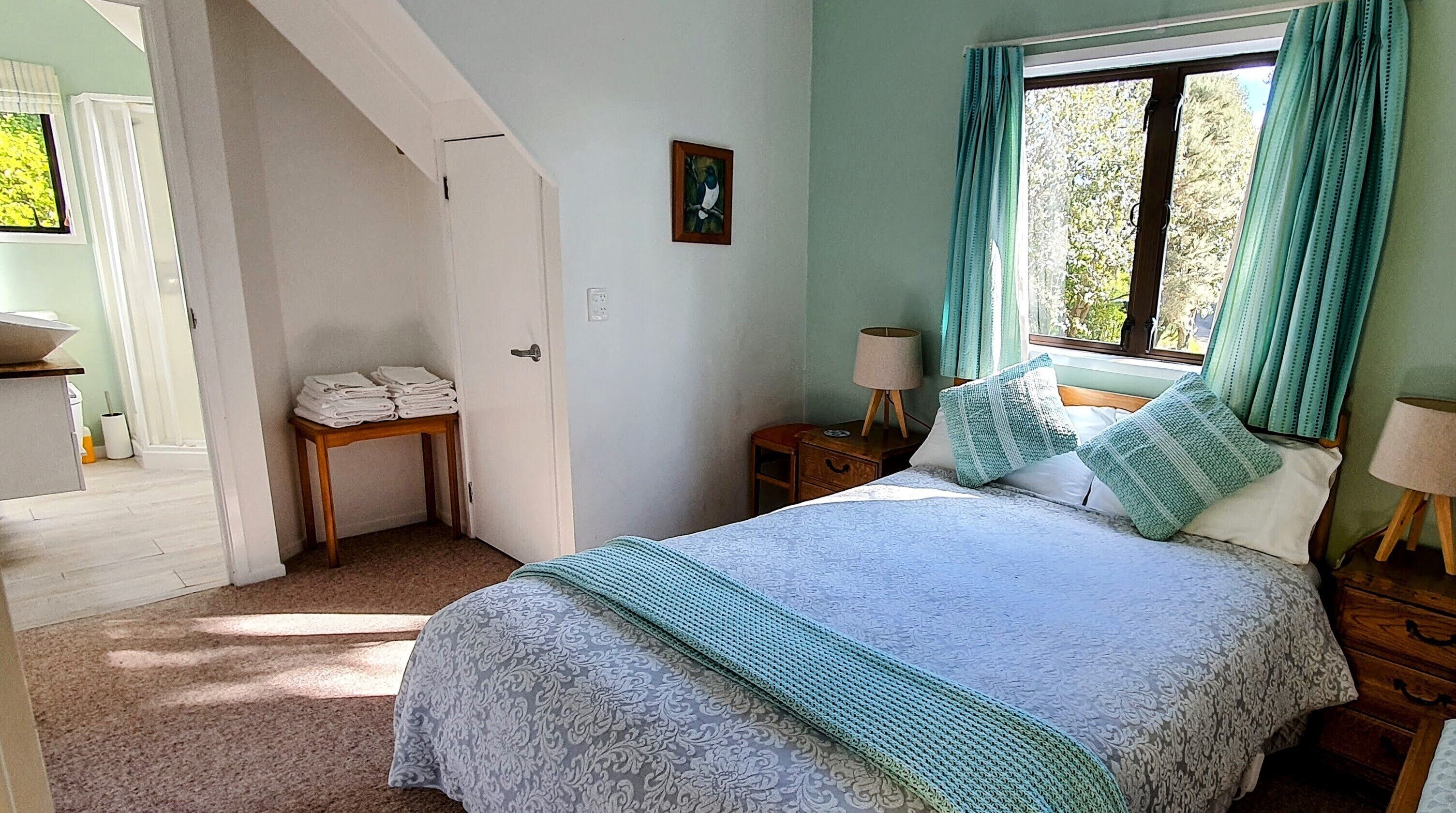 A bedroom with a bed, a bedside table with a lamp, a window with curtains, and a door leading to another room.