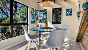 Un comedor con mesa y sillas de vidrio, ventanas amplias que dan a vistas de árboles y una lámpara colgante moderna.