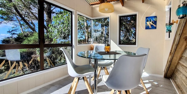 A dining area with a glass table and chairs, large windows offering a view of trees, and a modern pendant light.