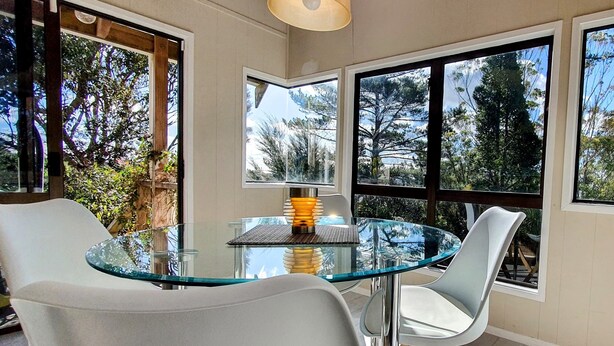Un comedor moderno con una mesa de vidrio y sillas blancas, ventanas amplias con vistas a árboles y una lámpara colgante.