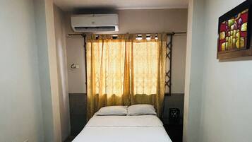 Een kleine, eenvoudige kamer met twee bedden, een raam met gordijnen, een airconditioning en een kunstwerk aan de muur.