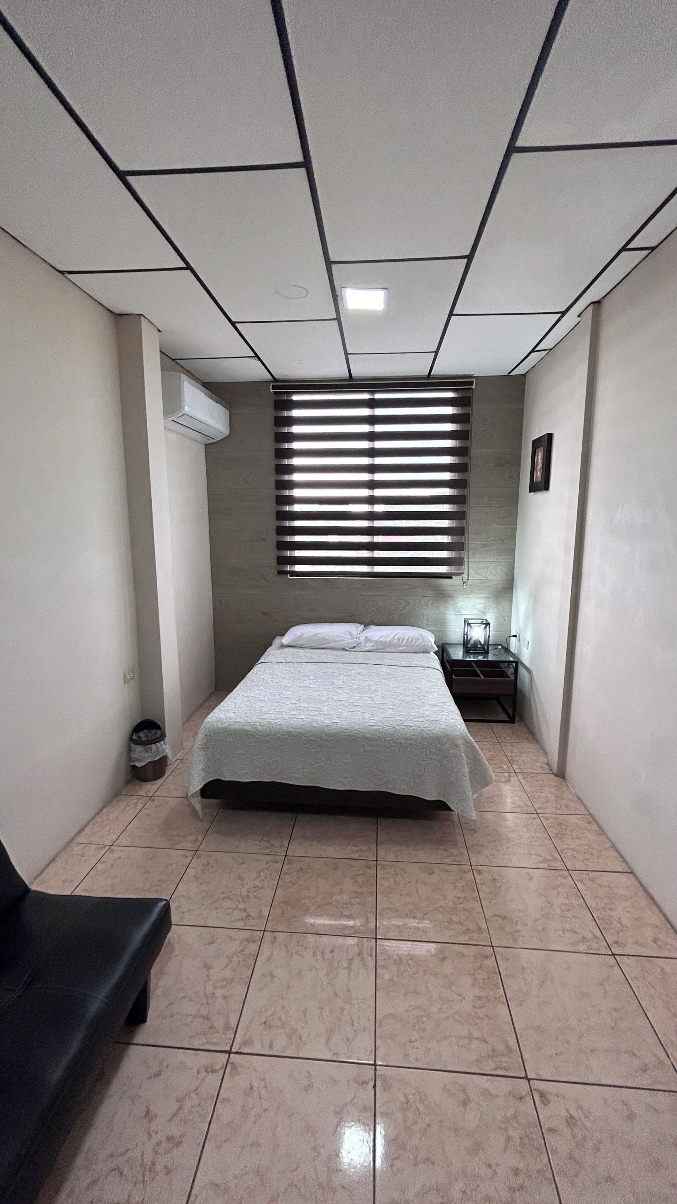 Een kleine, schone kamer met een bed, een stoel, een nachtkastje en een raam met jaloezieën.