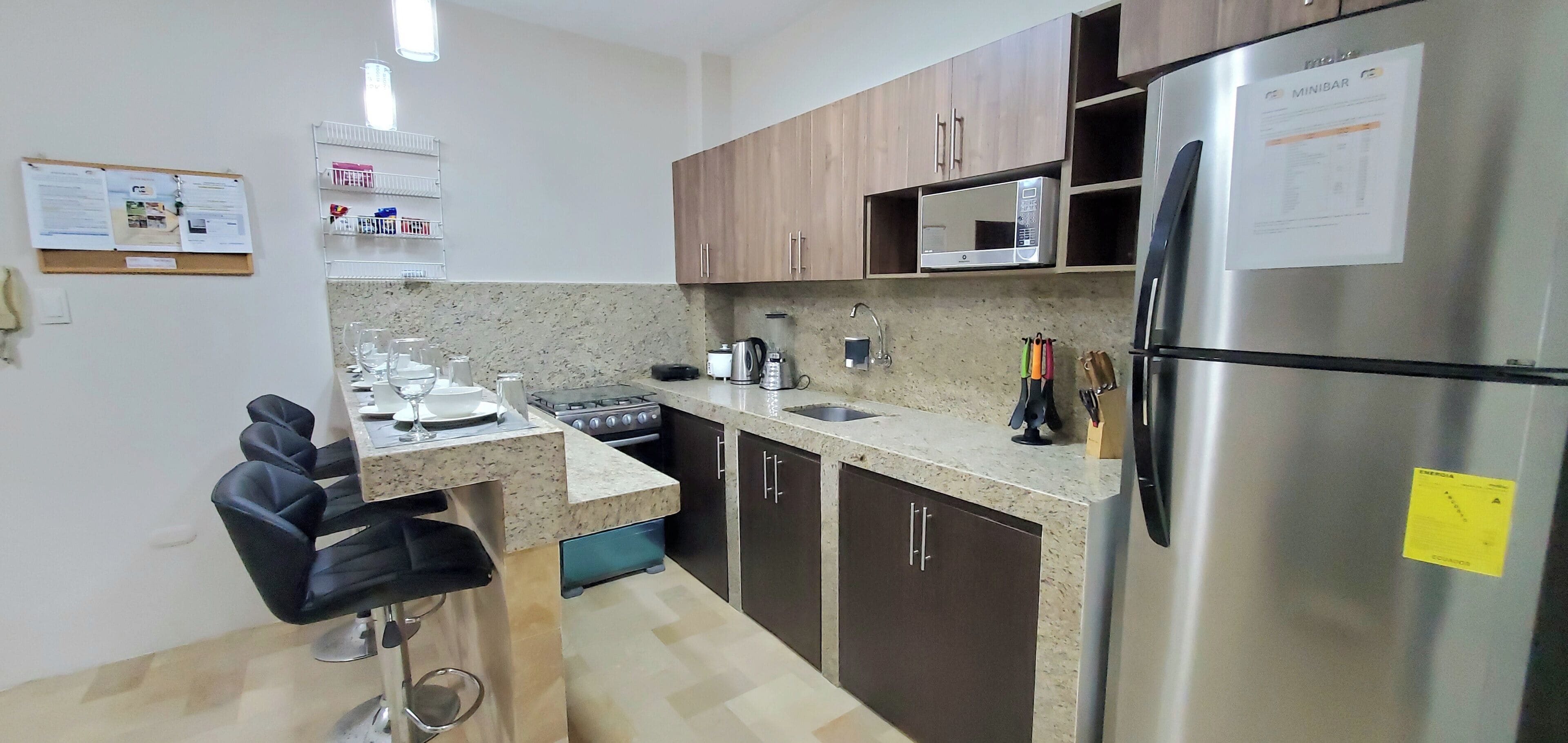 Cocina compacta con microondas integrado, refrigerador de acero inoxidable, estufa a gas y pileta. Hay taburetes de bar y una pequeña barra.