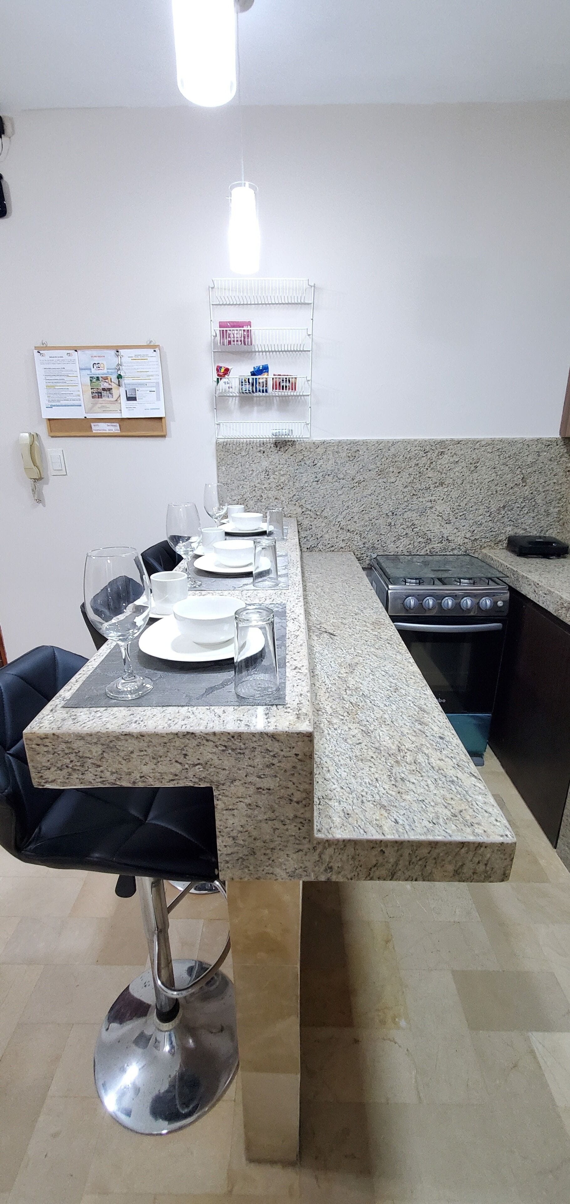 Een keuken met granieten aanrechtbladen, een gasfornuis, een magnetron en een koelkast. Er is een eettafel met stoelen en een prikbord aan de muur.
