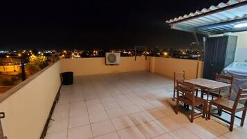 Uma varanda com mesa, cadeiras, uma lixeira e um aparelho de ar condicionado.