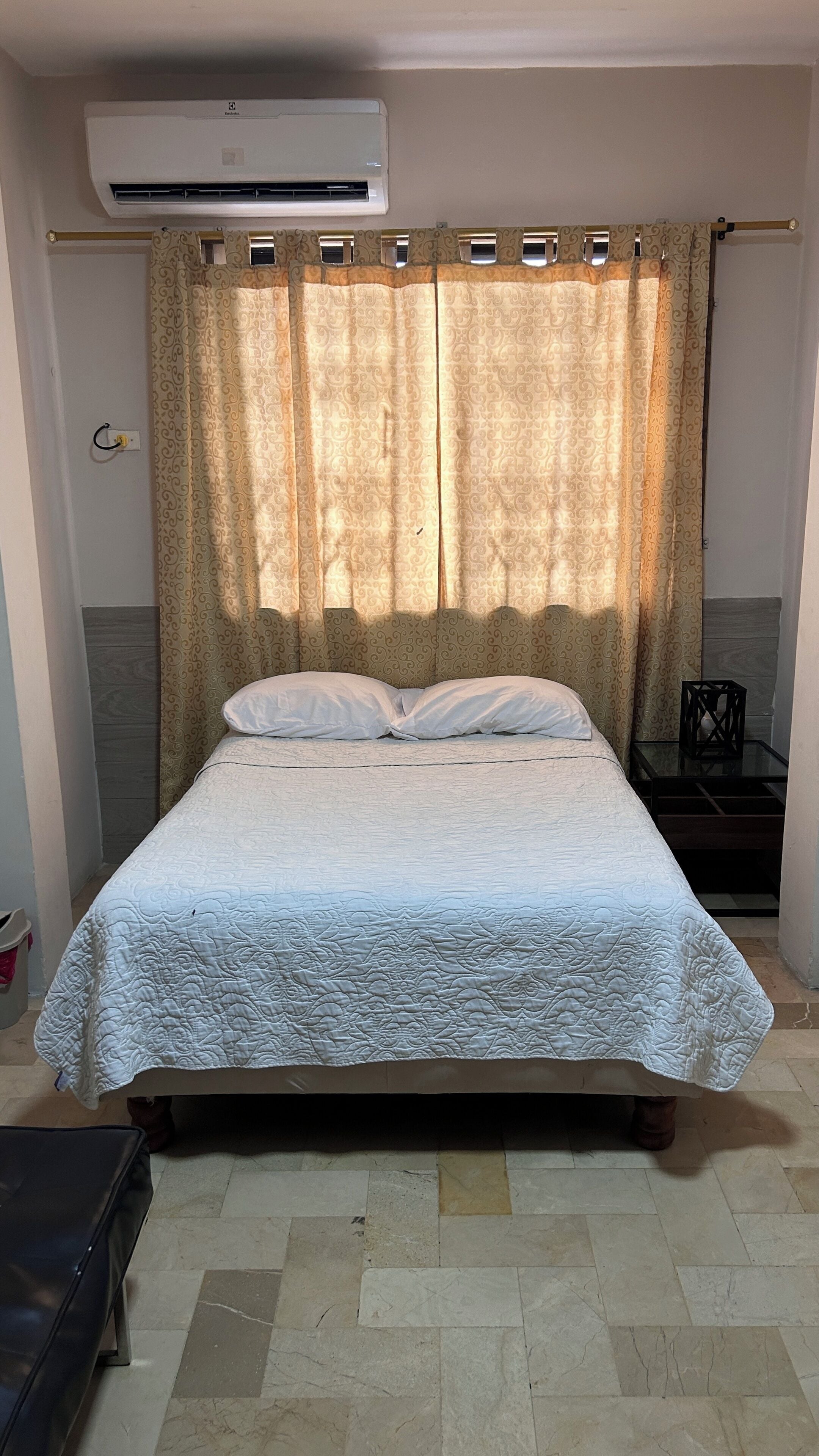 A bed with a patterned bedspread, a bedside table, an air conditioning unit, and a curtain with a paisley pattern.
