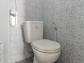 A toilet with a white seat and tank, a showerhead, and a tiled wall with a grid pattern. - Reddoorz Plus Near Taman Rasuna Menteng (Jakarta)