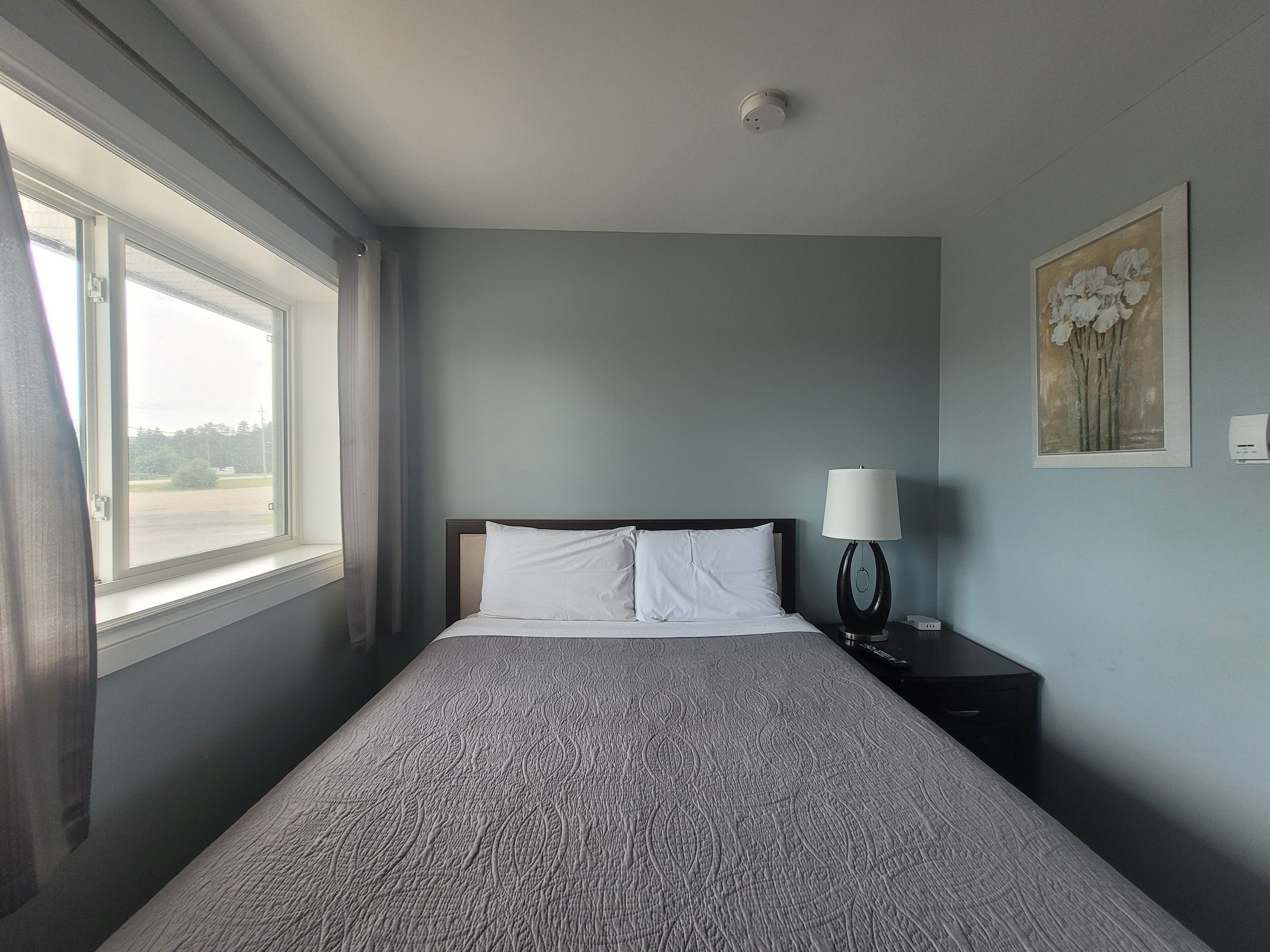 Une chambre à coucher avec un lit, une table de chevet, une lampe, une fenêtre et un tableau au mur.