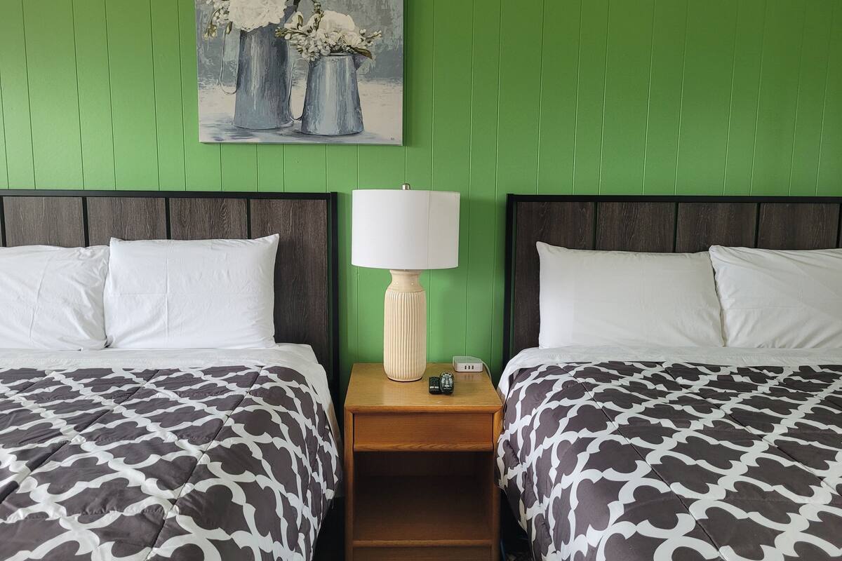 A hotel room with two beds, green walls, a painting of flowers, a bedside lamp, and a wooden nightstand.