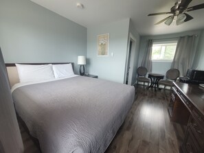 Quarto de hotel com uma cama grande, uma escrivaninha com cadeira, um ventilador de teto e uma janela com cortinas.