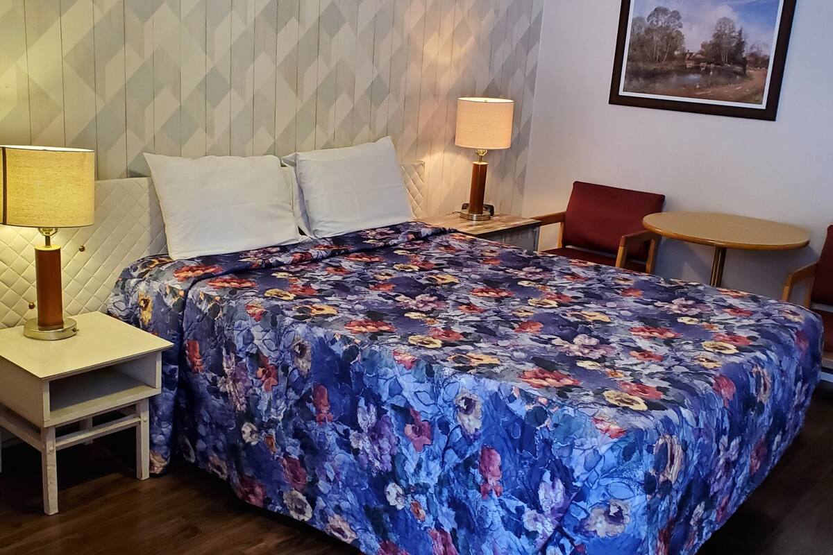 A hotel room with a bed, two bedside tables with lamps, a chair, a small table, a patterned rug, and a wall with a geometric design.