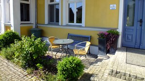 Een geel gebouw met een blauwe deur, een raam met een witte omlijsting, en een klein tafeltje met stoelen buiten.