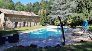 A wooden deck with a swimming pool, lounge chairs, and a stone building with shutters.