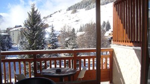 A balcony with a view of a snowy mountain and trees. There is a table with chairs and a small table lamp. - Studio accredits 3clés the slopes Centre station free wifi (Les Deux-Alpes)