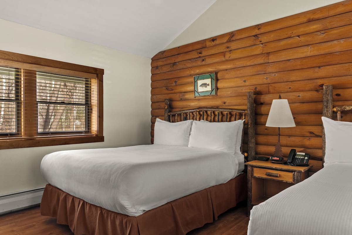 A room with a wooden log wall, a bed with white bedding, a bedside table with a lamp, and a framed picture on the wall.