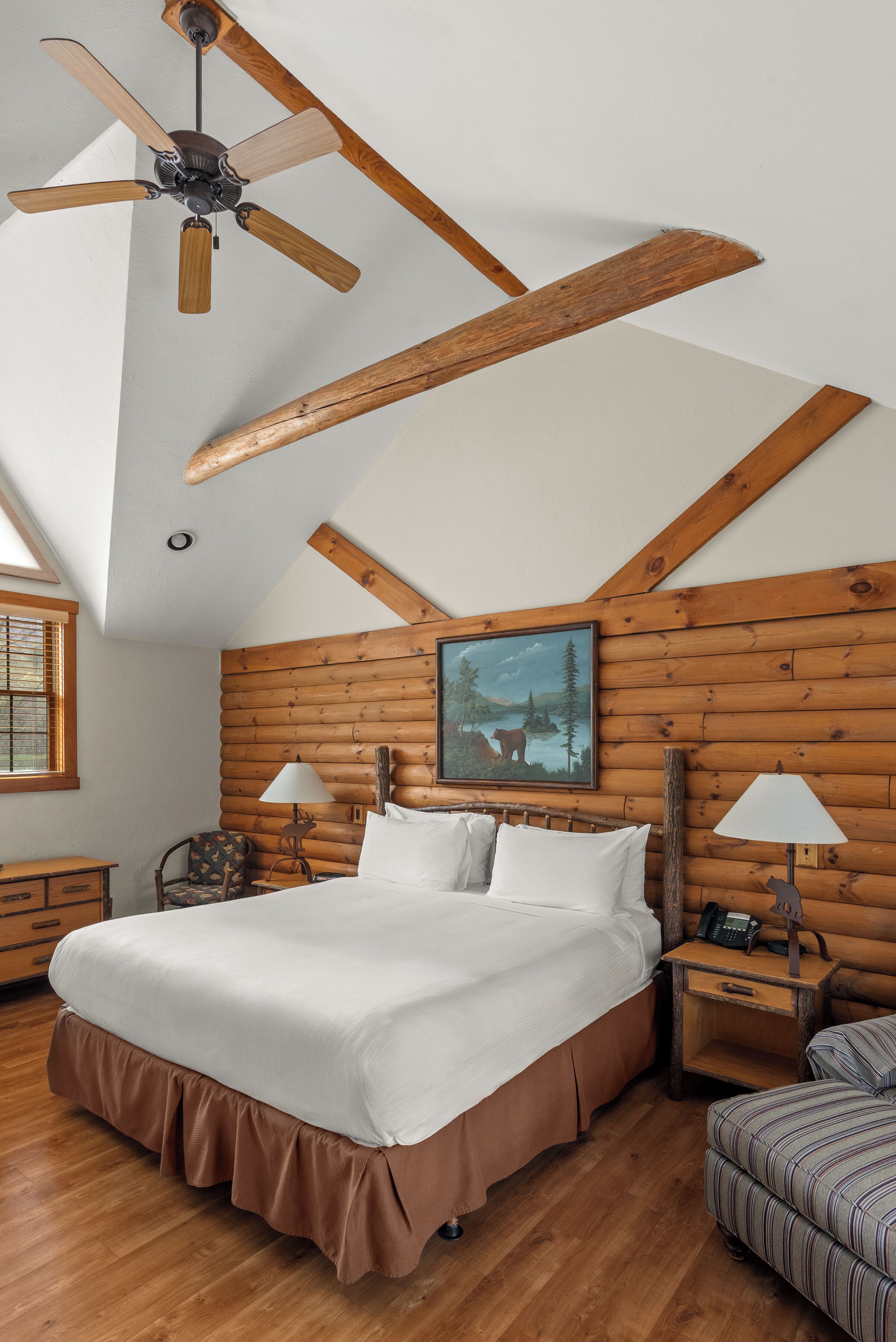 Una camera da letto con soffitto in legno, un letto, un comodino, una lampada, una sedia e un quadro appeso al muro.
