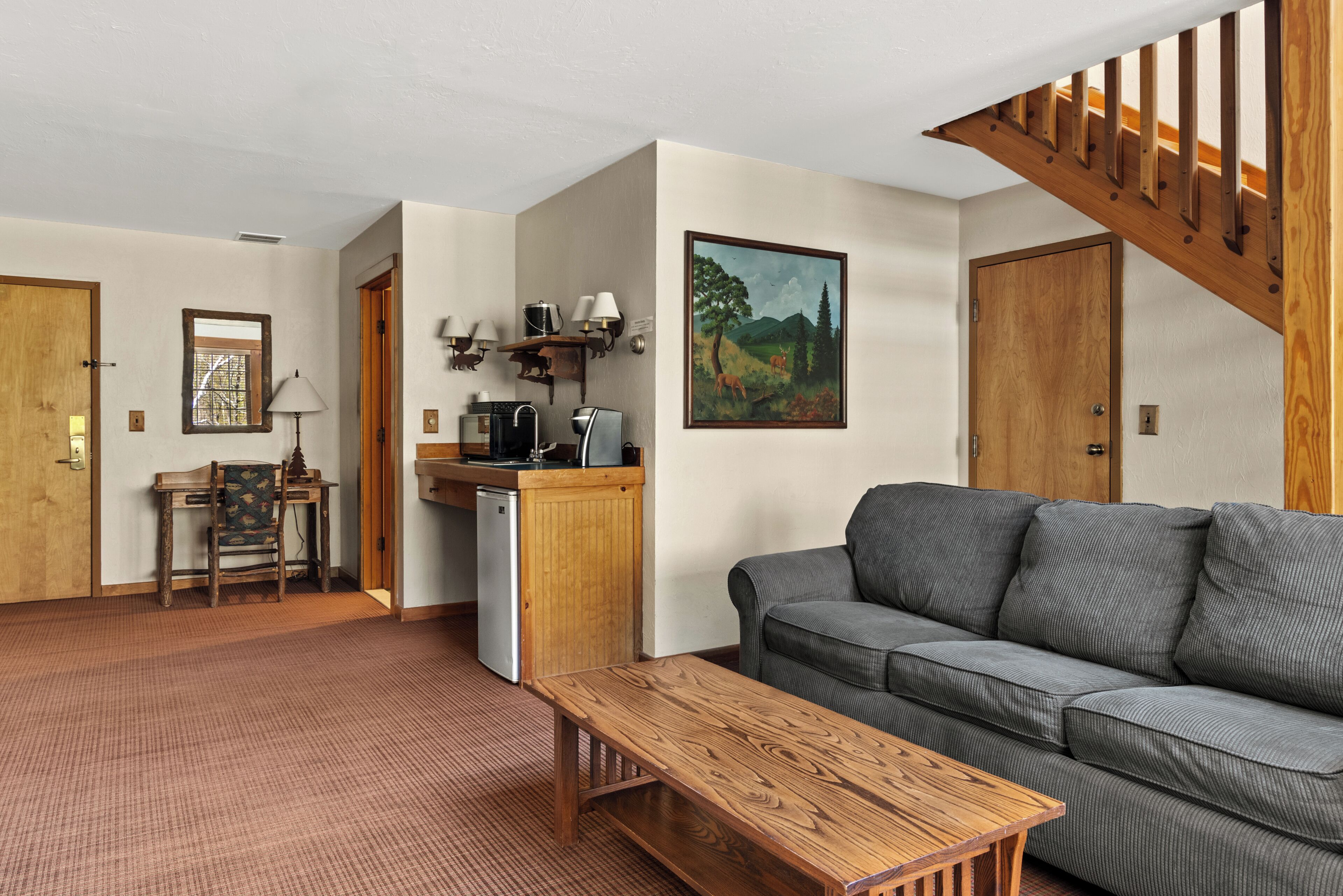 A living room with a sofa, a wooden table, a kitchen area with a refrigerator, a wooden staircase, and a painting on the wall.