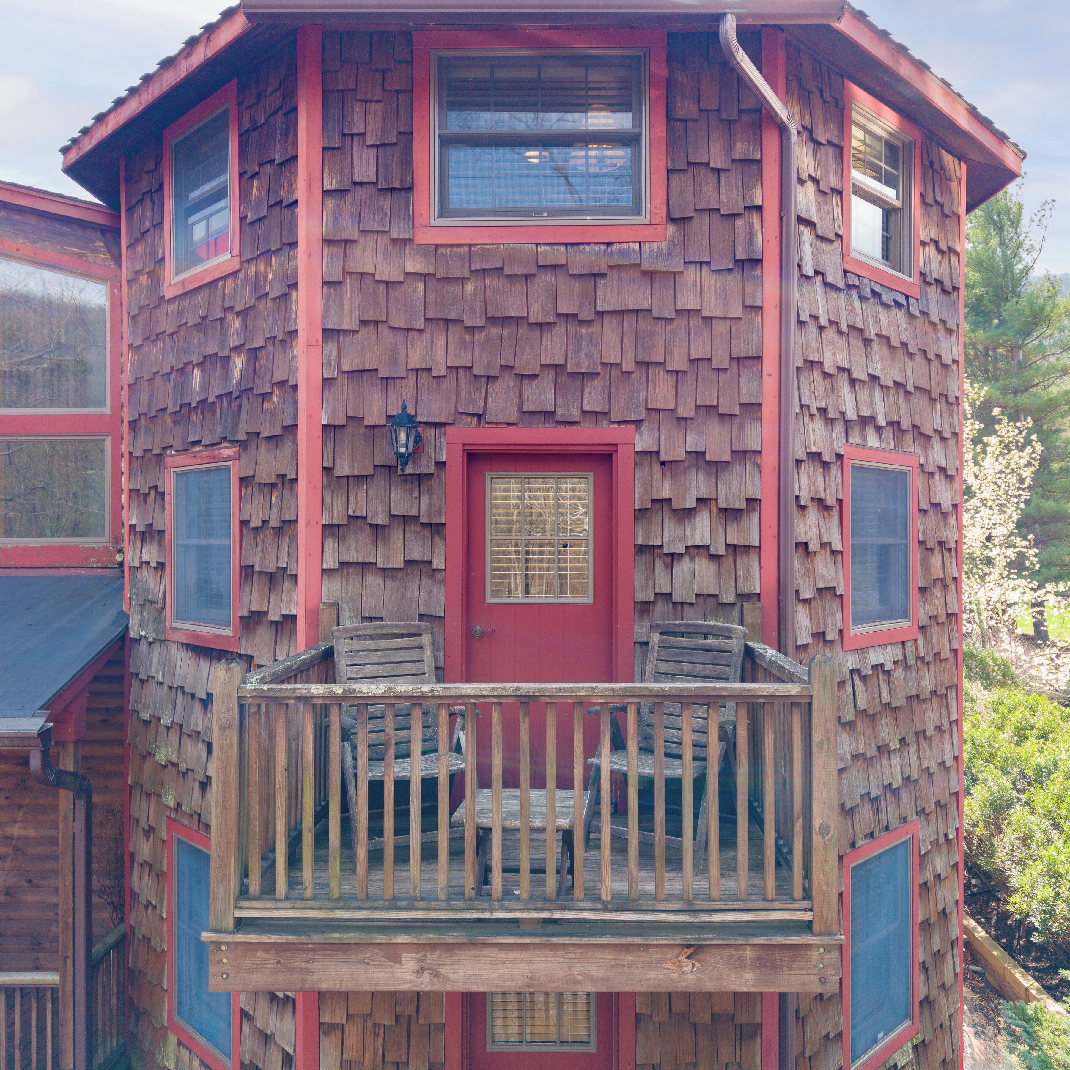A wooden cabin with a red door, multiple windows, and a balcony with chairs.