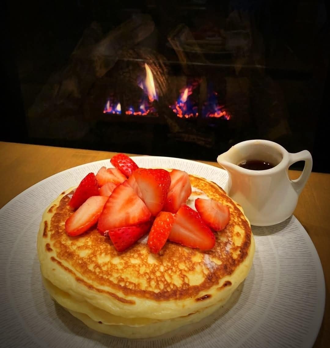 Una torre de panqueques cubiertos de fresas y una taza de café sobre una mesa con vista a un fuego.